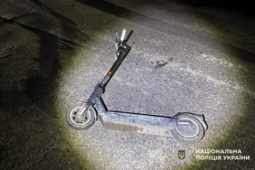 Electric scooter lying on its side on a dimly lit road at night, with the National Police of Ukraine logo in the corner.