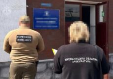 Two officials with blurred faces stand outside a brick building; one wears a Khmelnytskyi regional prosecutor's office jacket, the other a different uniform.