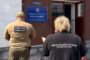 Two officials with blurred faces stand outside a brick building; one wears a Khmelnytskyi regional prosecutor's office jacket, the other a different uniform.