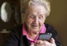 Smiling elderly woman in a pink collared shirt looks at a smartphone in her hands at home.