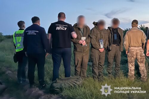 Group of uniformed officers standing in a field with blurred detainees, some in camouflage, in a police operation scene; Ukrainian National Police logo visible on the bottom right.