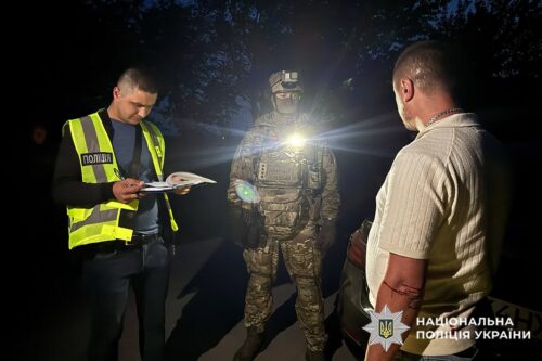 Nighttime scene of a Ukrainian police officer in a reflective vest taking notes, a camouflaged soldier with a flashlight stands between him and a man with a tattooed arm; National Police of Ukraine logo visible.