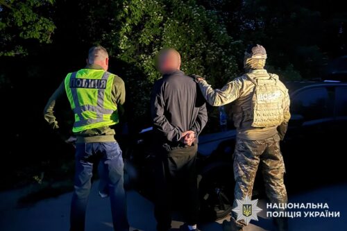 Two police officers escorting a handcuffed man beside a car during an arrest; National Police of Ukraine emblem visible in the corner.