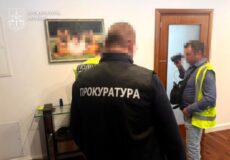 Two men in a room; one wearing a black jacket labeled 'ПРОКУРАТУРА' with blurred faces; another man in a yellow vest with a camera.