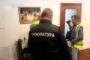 Two men in a room; one wearing a black jacket labeled 'ПРОКУРАТУРА' with blurred faces; another man in a yellow vest with a camera.