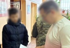 Three men stand in a hallway; one in a light pink shirt holds a sheet of paper while two others listen; faces are blurred, with a Ukrainian prosecutor's office sign visible in the background.