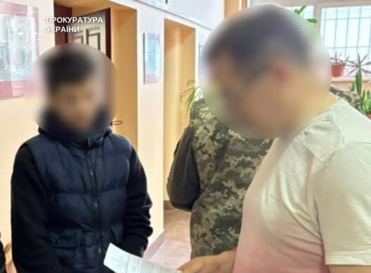 Three men stand in a hallway; one in a light pink shirt holds a sheet of paper while two others listen; faces are blurred, with a Ukrainian prosecutor's office sign visible in the background.