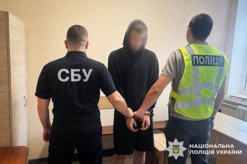 Suspect in a black hoodie is handcuffed by two officers—one in a dark SBU shirt and one in a reflective police vest—in a small room.