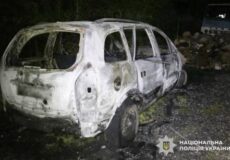 Burned-out white hatchback car at night, severely damaged by fire, with Ukrainian police insignia in the lower corner.