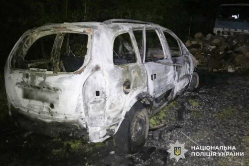 Burned-out white hatchback car at night, severely damaged by fire, with Ukrainian police insignia in the lower corner.