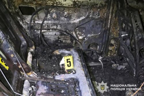 Inside a burned car engine bay with charred wiring and debris; evidence marker '5' placed on a metal surface, with National Police of Ukraine logo in the corner.