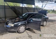 Black sedan under a metal carport with the hood up and the driver’s door open, engine visible in the bay area on a gravel surface while a second door is also ajar.