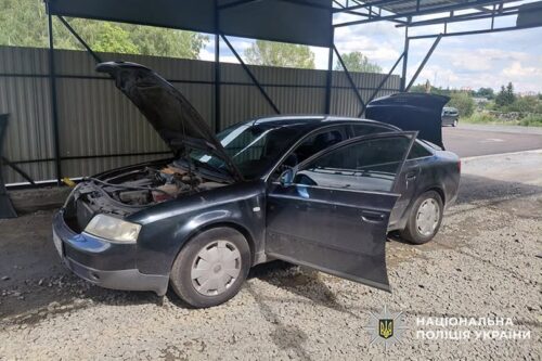 Black sedan under a metal carport with the hood up and the driver’s door open, engine visible in the bay area on a gravel surface while a second door is also ajar.
