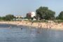 Crowded lakeside beach with swimmers and sunbathers; trees and buildings in the background.