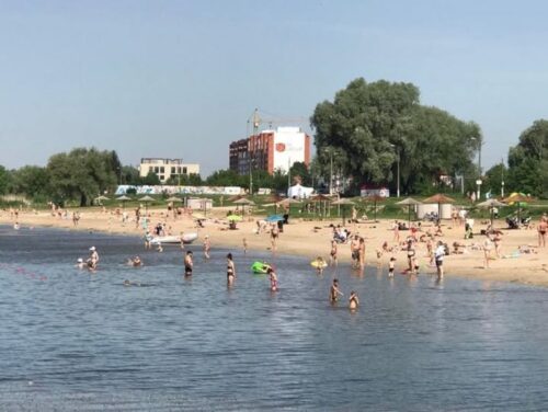 Crowded lakeside beach with swimmers and sunbathers; trees and buildings in the background.