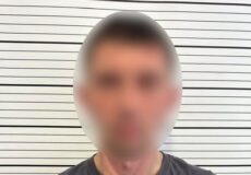 Mugshot-style photo of a man with blurred face, wearing a dark shirt with Ukrainian National Police logo, standing against a striped height chart.