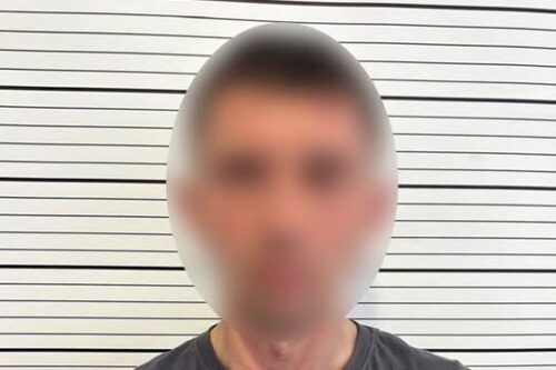 Mugshot-style photo of a man with blurred face, wearing a dark shirt with Ukrainian National Police logo, standing against a striped height chart.