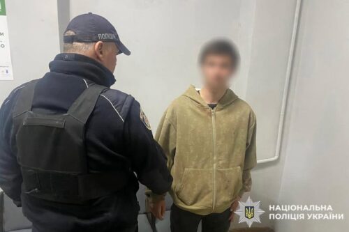 Ukrainian police officer in a dark uniform speaks to a young man in a tan hoodie inside a facility; police watermark visible.