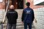 Two investigators, one in a black jacket and the other in a dark blue vest labeled with Ukrainian prosecutor and DBR insignia, walk up concrete steps toward a brick entrance.