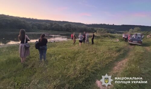 Group of people on a grassy riverbank at sunset, with parked cars; Ukrainian police watermark in the bottom right.