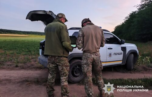 Two officers in camouflage stand beside a white Ukrainian police SUV with the trunk open in a rural field.