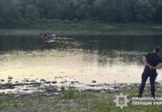 Man stands on a grassy riverbank with hands behind his back, watching a small boat with people in the water nearby; Ukrainian National Police watermark in the corner.