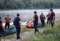 Rescue team in life jackets gathers by inflatable boats on a lakeshore, with a crew member carrying an orange container near the water.
