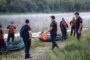 Rescue team in life jackets gathers by inflatable boats on a lakeshore, with a crew member carrying an orange container near the water.