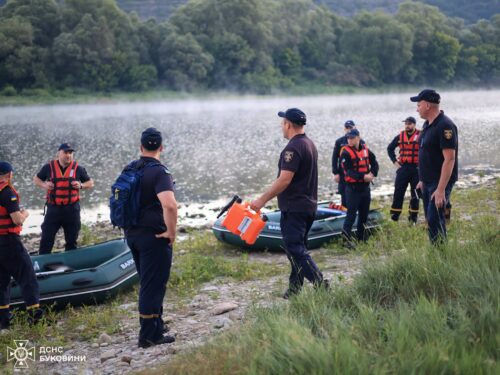 Rescue team in life jackets gathers by inflatable boats on a lakeshore, with a crew member carrying an orange container near the water.