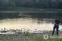 Man stands on a grassy riverbank with hands behind his back, watching a small boat with people in the water nearby; Ukrainian National Police watermark in the corner.