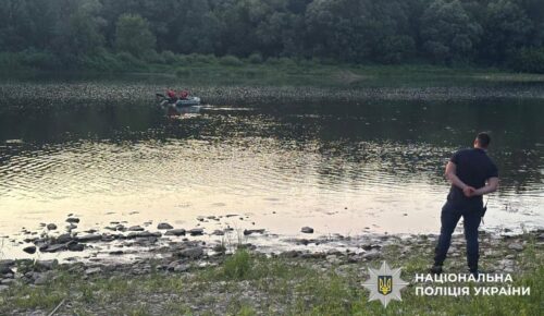Man stands on a grassy riverbank with hands behind his back, watching a small boat with people in the water nearby; Ukrainian National Police watermark in the corner.