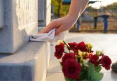Person's hand wipes a stone monument with a cloth; red roses lie nearby at its base.