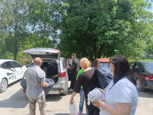 Group of people loading bags from the open trunk of a silver SUV in a sunny parking lot with green trees in the background.
