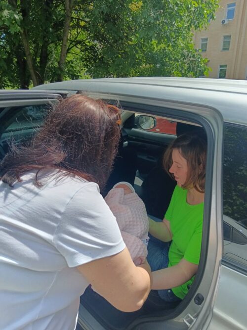 Two women help place a baby wrapped in pink into a car seat inside a silver car, with one woman inside and another outside the vehicle.