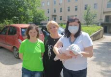 Three women stand outdoors near an apartment building; one woman in a white shirt holds a newborn wrapped in a blanket, wearing a mask.