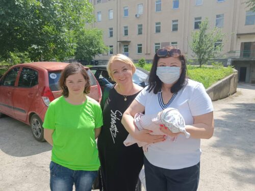 Three women stand outdoors near an apartment building; one woman in a white shirt holds a newborn wrapped in a blanket, wearing a mask.