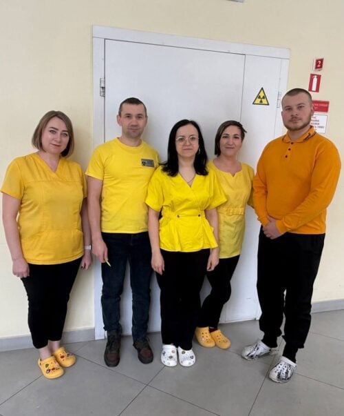 Five staff members in yellow and orange scrubs pose indoors in front of a door with a radiation symbol.