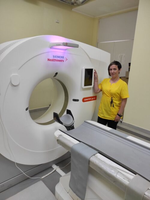 Healthcare worker in a yellow shirt stands beside a large Siemens Healthineers CT scanner in a hospital room, patient table ready for scanning.
