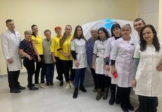 Group of doctors and nurses in white coats posing in a clinic, with a medical imaging device visible in the background.