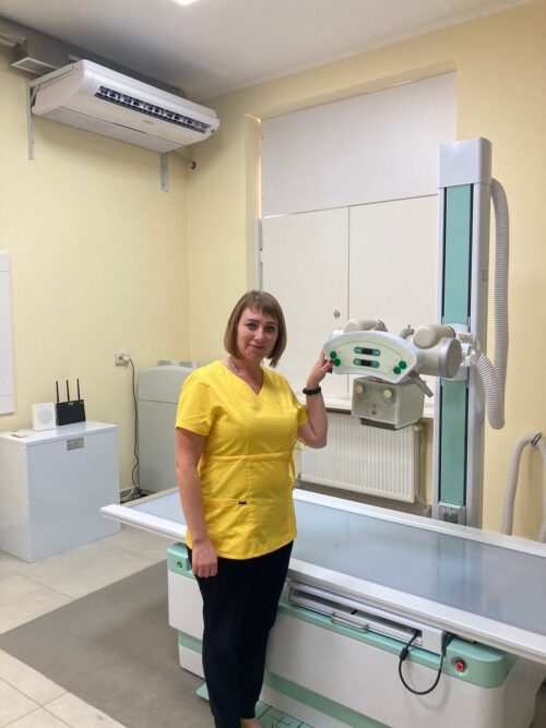 A woman in a yellow medical uniform stands beside an X-ray machine in a radiology room, smiling at the camera.
