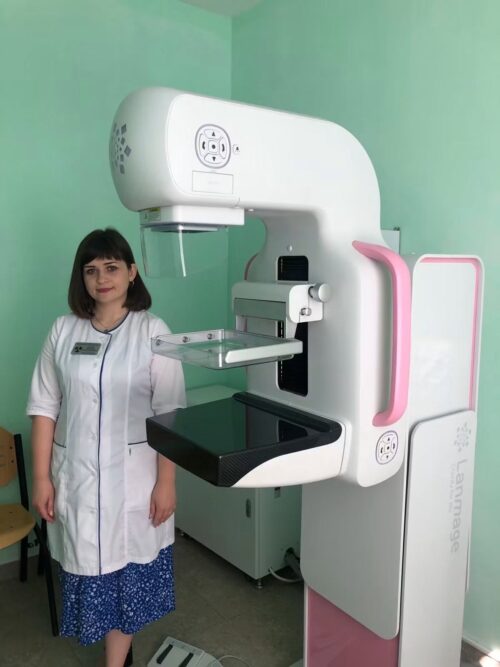 Female healthcare worker in a white coat stands beside a large white medical imaging machine with a pink accent panel in a clinic room.