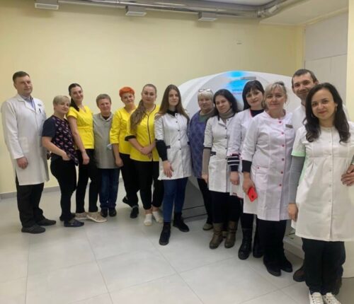 Group of doctors and nurses in white coats posing in a clinic, with a medical imaging device visible in the background.