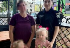 Ukrainian policewoman in uniform posing outdoors with two women and two young girls, all standing beside a decorative metal fence; some faces pixelated for privacy.