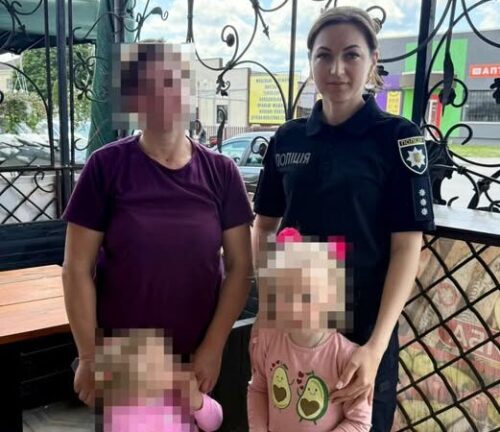 Ukrainian policewoman in uniform posing outdoors with two women and two young girls, all standing beside a decorative metal fence; some faces pixelated for privacy.