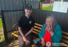 Young Ukrainian police officer sits on a bench beside an elderly woman in a teal cardigan who is smoking outside, with police emblem visible on the woman's badge.