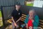 Young Ukrainian police officer sits on a bench beside an elderly woman in a teal cardigan who is smoking outside, with police emblem visible on the woman's badge.