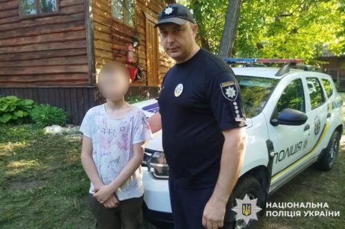 Policeman in uniform stands beside a young girl with blurred face; a Ukrainian police SUV is parked behind them near a wooden cabin.