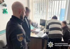 Two men stand at a desk with computers as officers assist civilians in a small office; Ukrainian National Police logo visible in corner