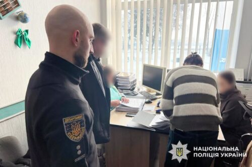 Two men stand at a desk with computers as officers assist civilians in a small office; Ukrainian National Police logo visible in corner