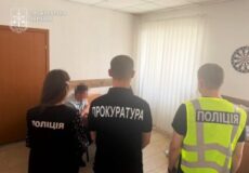 Three officers stand inside a room: a prosecutor in a black shirt reading 'ПРОКУРАТУРА' faces away, flanked by a police officer in a black 'Поліція' shirt and another officer in a bright vest marked 'Поліція'. A blurred person is present near them.
